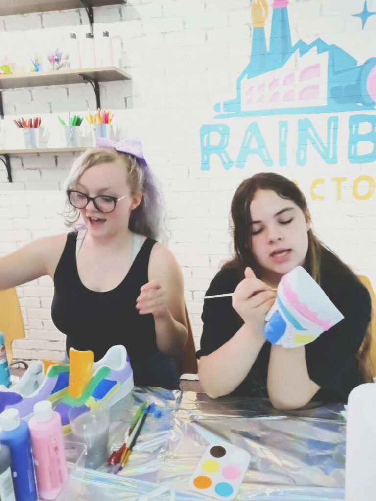 Two girls sitting at a table with paint and paint brushes.