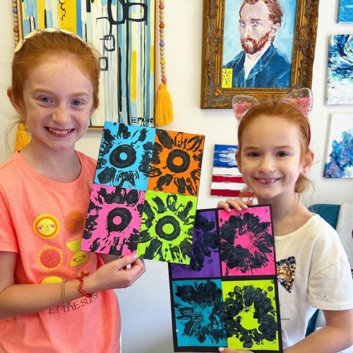 Two girls holding up colorful paintings in an art studio.