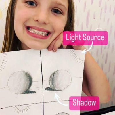 A little girl holding up a drawing of a light source and shadow.